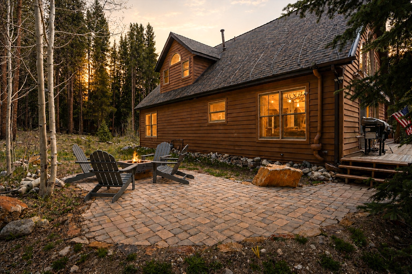 Cabin at dusk with paver patio, fire pit, and Adirondack chairs under glowing windows