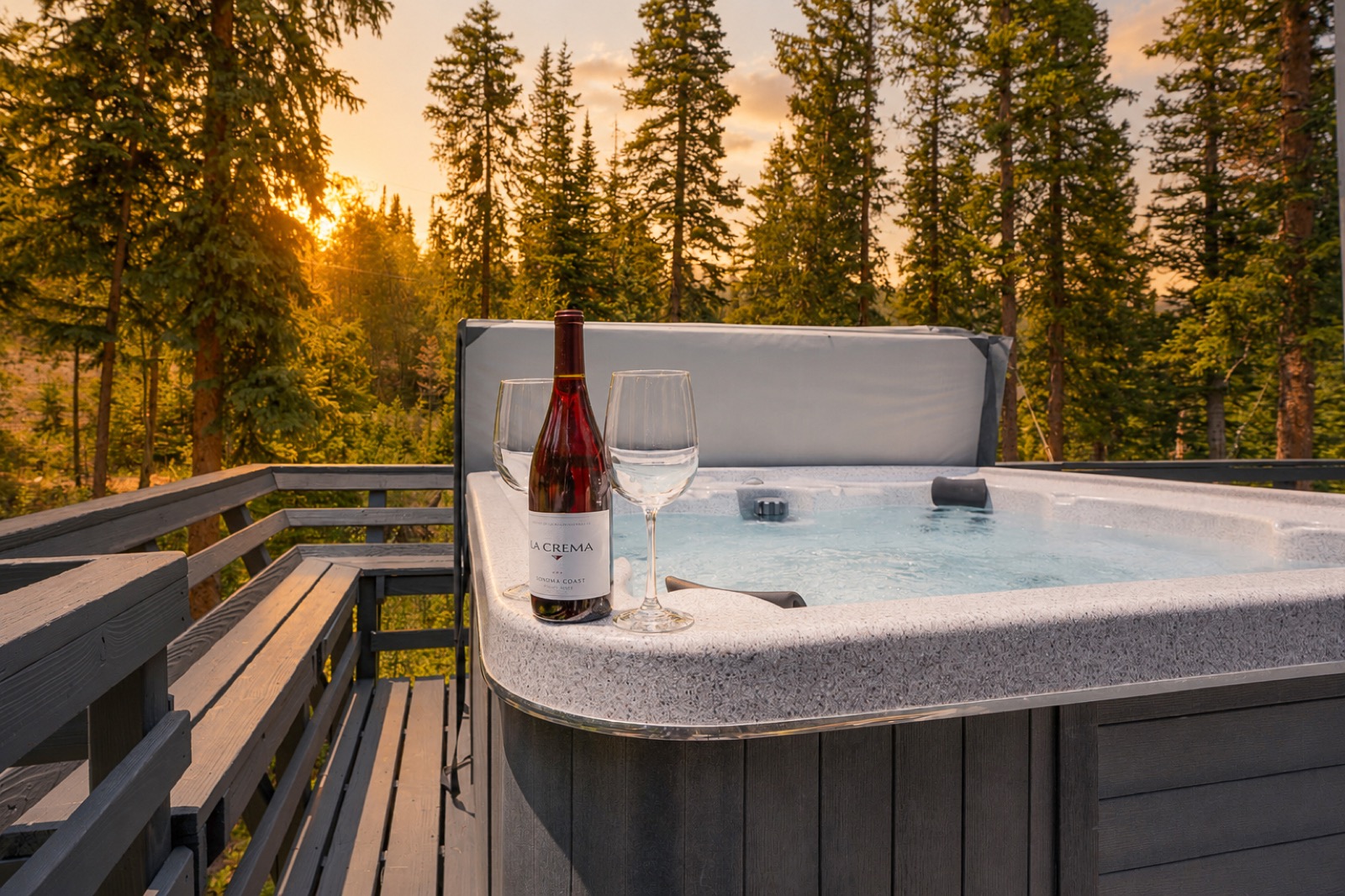 Wine bottle and glasses on the edge of a hot tub at sunset with pines behind