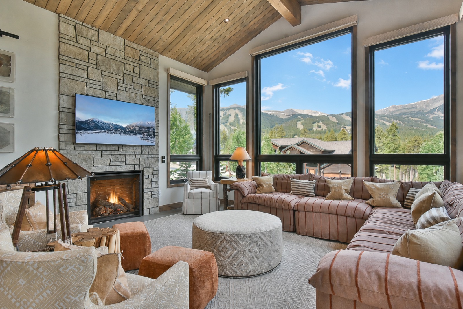 Luxury mountain living room with stone fireplace, vaulted wood ceiling, and floor-to-ceiling windows framing the ski slopes